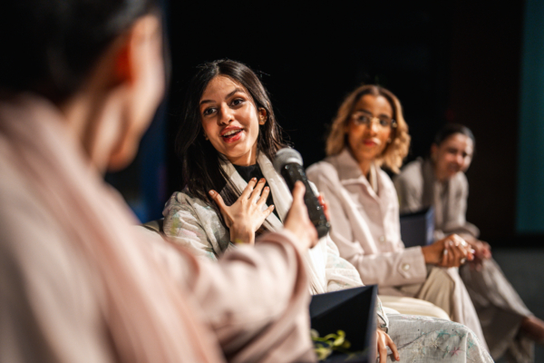 Middle Eastern woman panelist engaging audience while speaking into microphone during inspiring university event in Riyadh auditorium, with attentive listeners in background